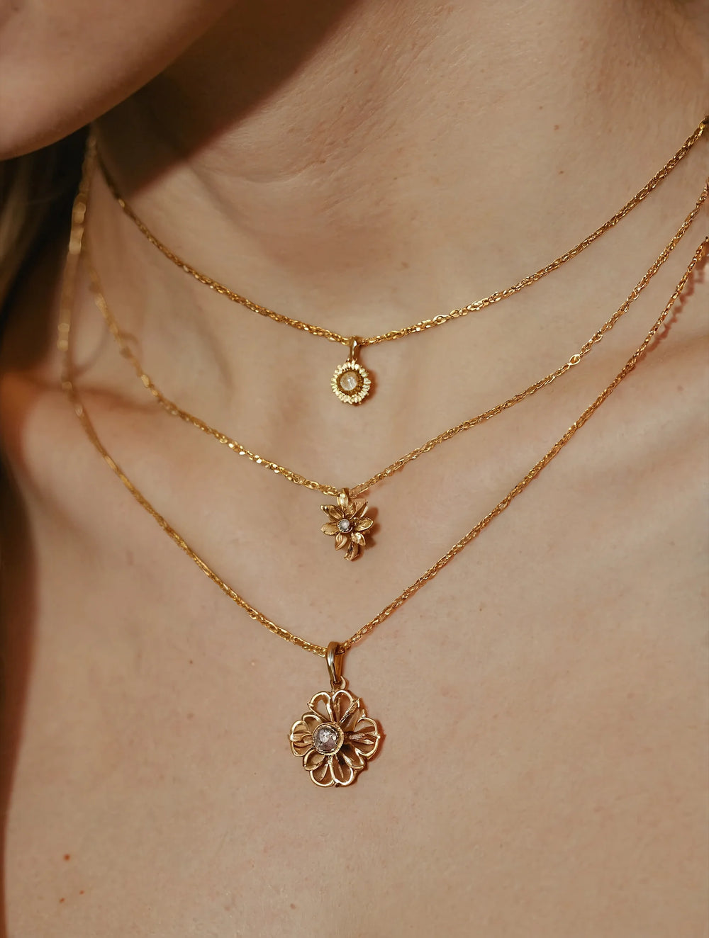 Close-up of a person wearing two gold necklaces with floral pendants on a neutral background