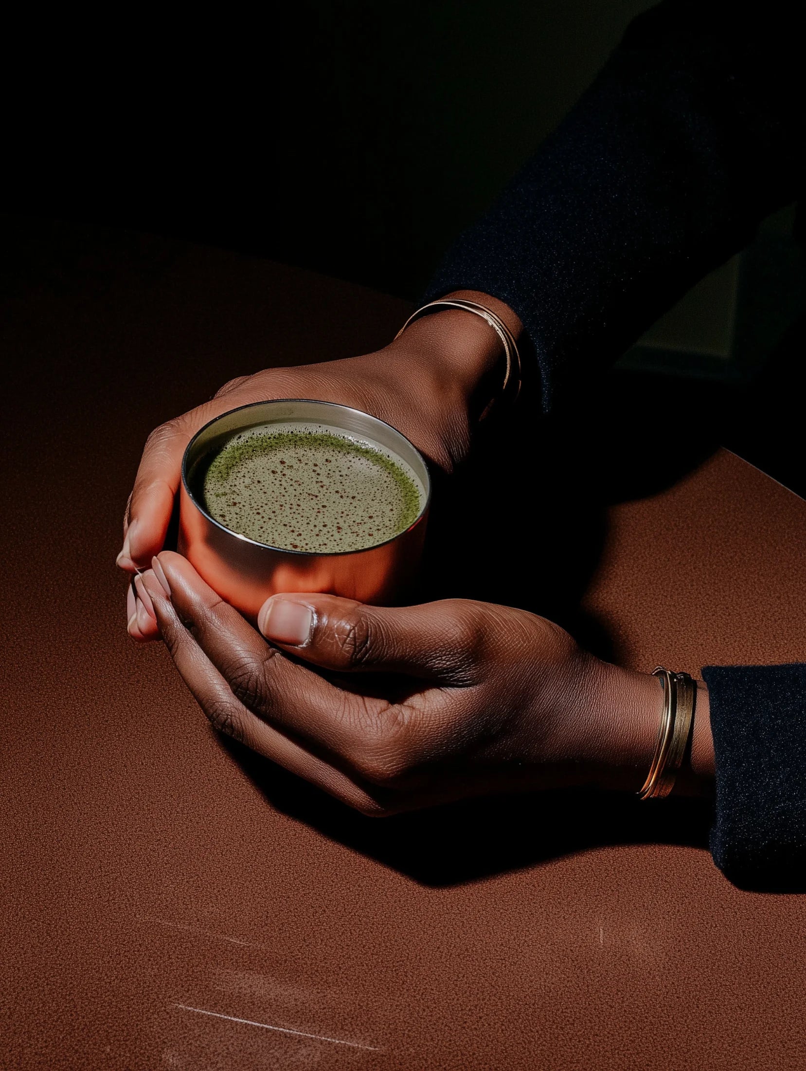 Hands holding a small metallic cup with matcha on a dark surface