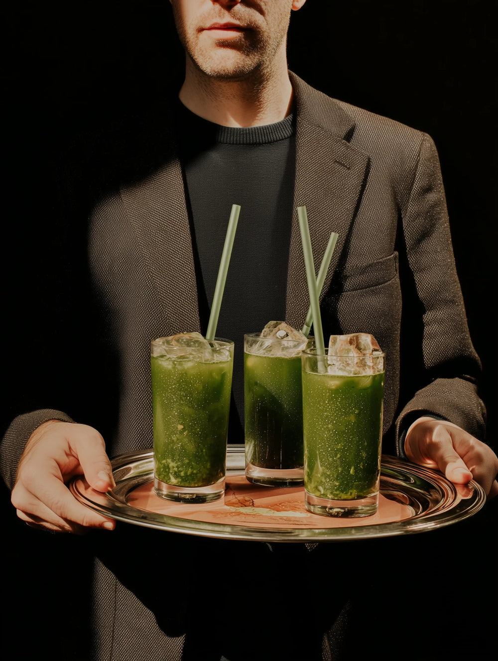 Man holding a tray with three green smoothies and straws against a dark background