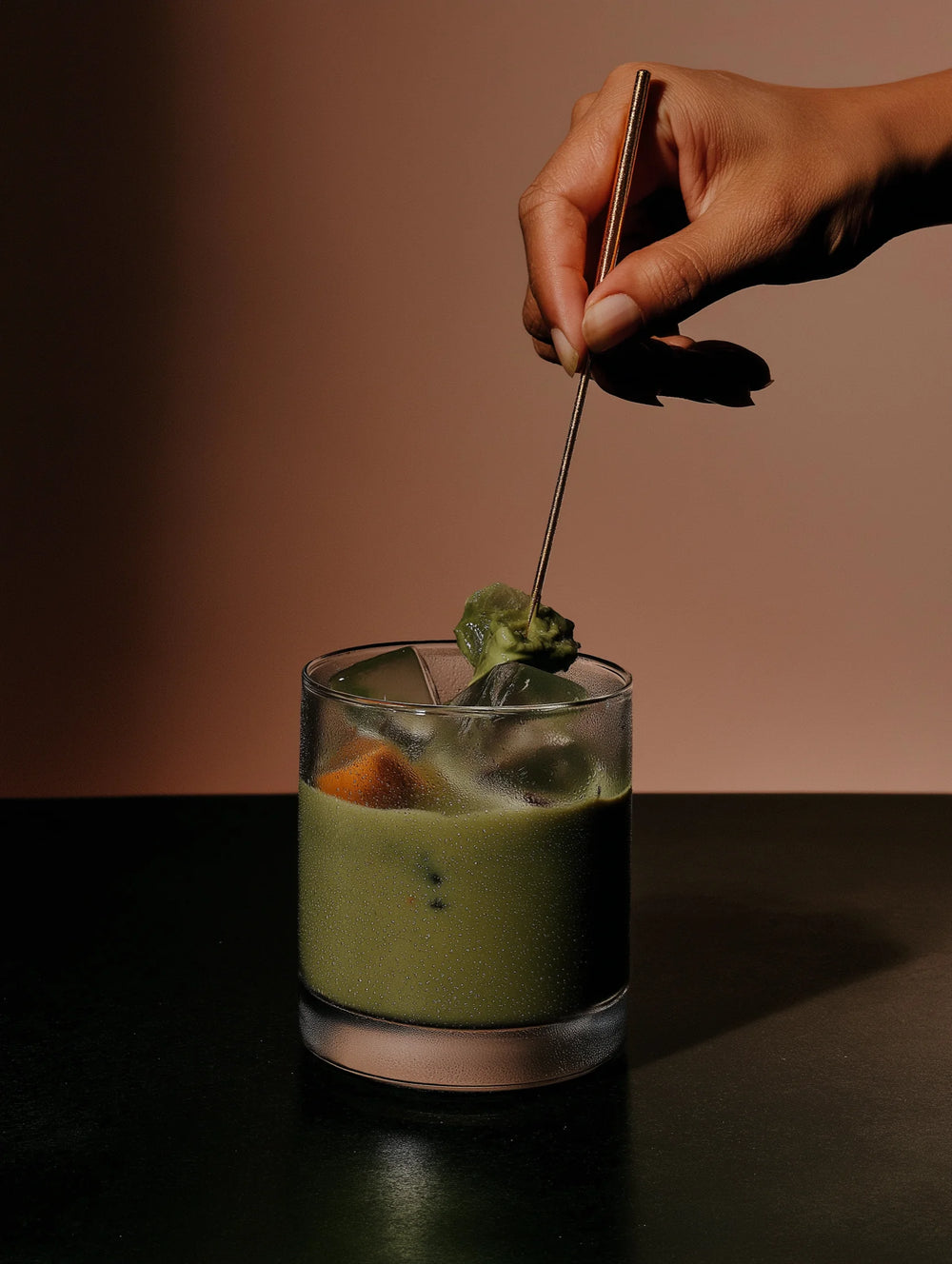 Hand stirring a green matcha in a glass with a spoon against a brown background