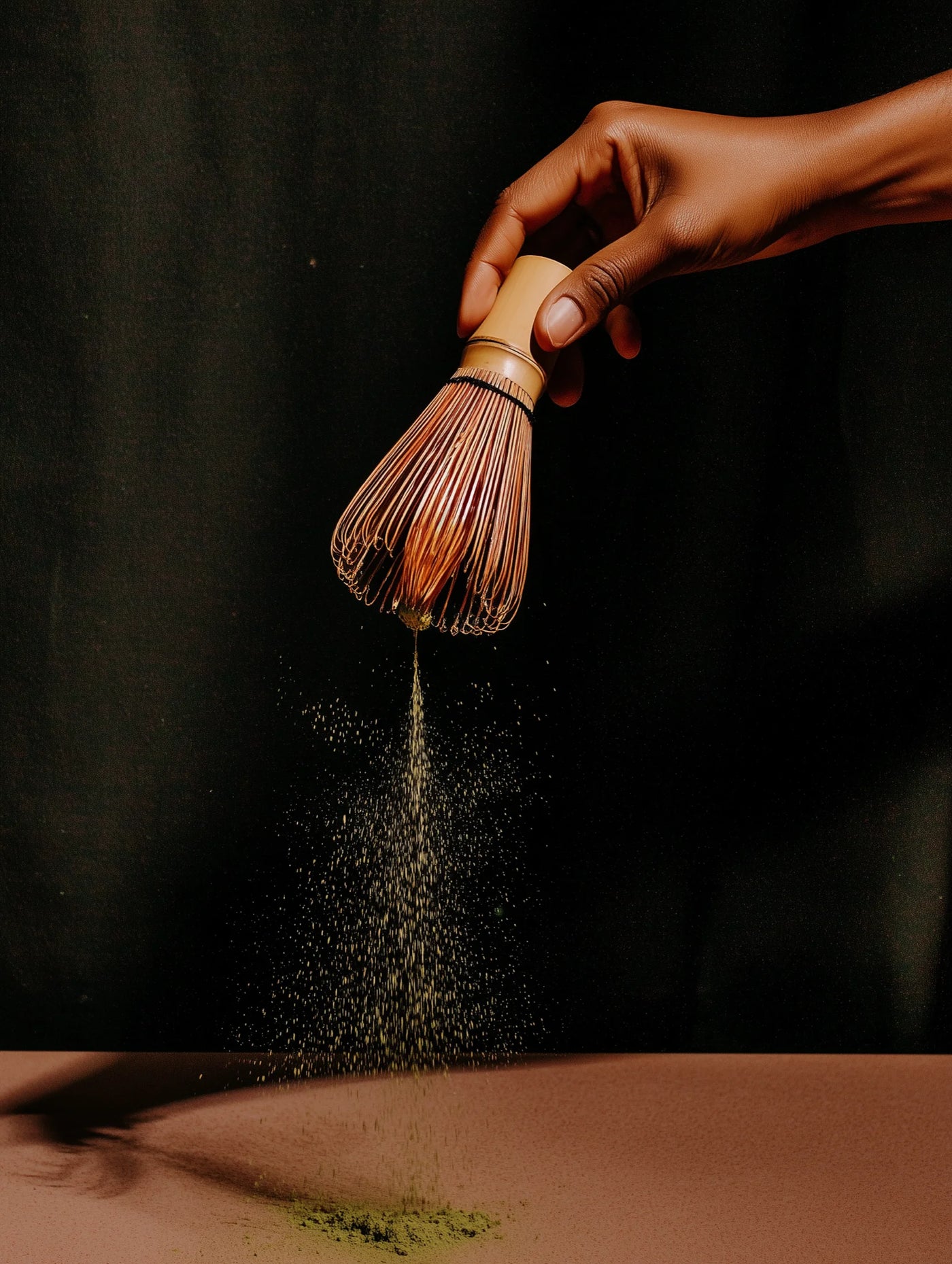 Hand using a wooden whisk to sprinkle matcha powder on a dark background